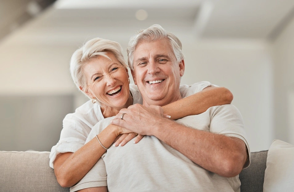 Happy older couple laughing and smiling together in a bright living room — confident, natural-looking smiles, MAP Dental Center Houston West Houston.